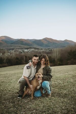 Smiling couple crouching with golden retriever dog in mountain meadow landscape.