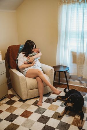 Young mother sitting in armchair cradling newborn baby while black and tan dog rests on checkered rug nearby.