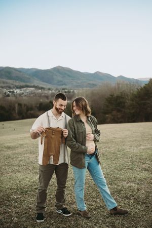 Happy couple holding baby outfit outdoors with mountain backdrop during maternity shoot.