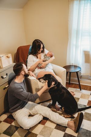 A young family with a newborn baby, father petting a black dog on a checkered rug in a cozy nursery room.