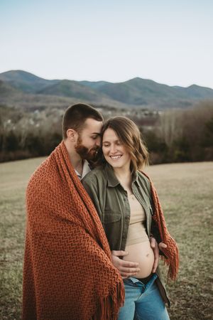 Couple wrapped in rust knit blanket embracing outdoors showing pregnant belly in mountains.