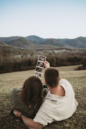 Couple sitting on grassy hillside viewing ultrasound photos together with mountain range backdrop.