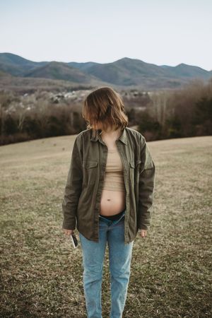 Pregnant woman in olive jacket and jeans showing baby bump in open field with mountains behind her.