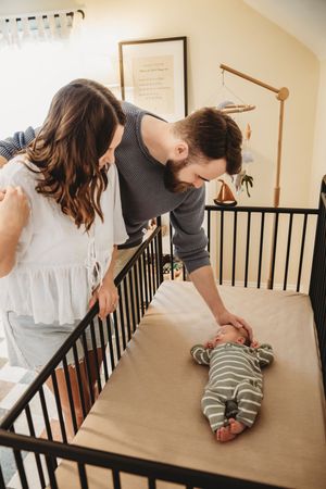 Parents lovingly lean over a dark wood crib to check on their newborn baby in a cozy nursery room.