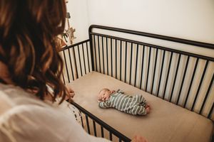 Mother gazing lovingly at newborn baby sleeping peacefully in a black metal crib on a beige fitted sheet.