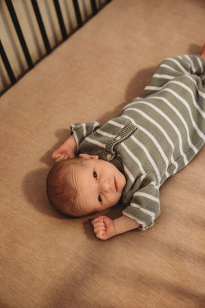 A newborn baby in a green striped outfit lies awake in a wooden crib on a beige sheet.