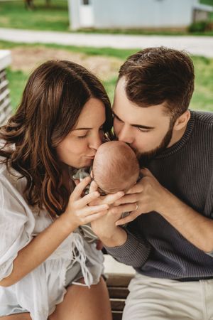 Young parents kissing their newborn baby outdoors, a tender family moment captured in natural light.