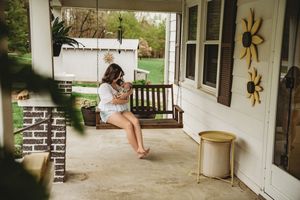 Woman sitting on a wooden porch swing holding a small dog outside a charming white farmhouse.