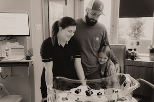 Black and white photo of parents and young child admiring newborn baby in hospital bassinet.
