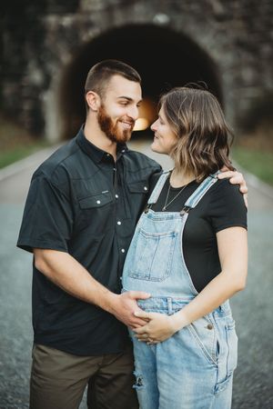 Smiling couple embracing on a road in front of a tunnel, woman in overalls cradling her baby bump.