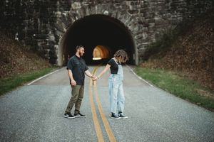 Couple holding hands on a road in front of a stone tunnel arch, standing on yellow center lines.