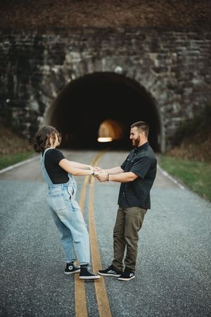 Couple holding hands on a road in front of a stone tunnel, woman in overalls and man in black shirt.