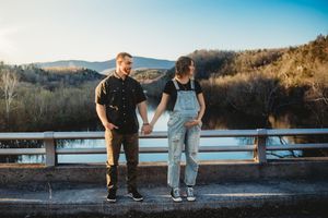 Couple holding hands on a bridge wearing casual outfits with mountain and river backdrop at sunset.