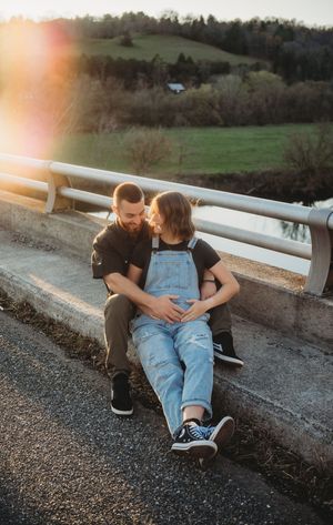Couple sitting on a bridge curb at sunset, tenderly cradling a pregnant belly in overalls.