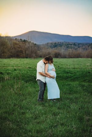 Couple embracing in a green field at sunset, gently holding a pregnant belly with mountains behind.