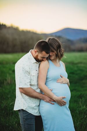 Couple sharing a tender moment in a green field at sunset, woman in blue dress showing pregnancy bump.
