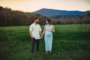 Couple holding hands in green field at sunset with mountain backdrop, woman in blue maternity dress.