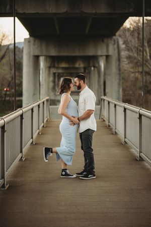 Pregnant couple sharing a tender moment on a bridge, woman in a light blue dress lifting one foot.