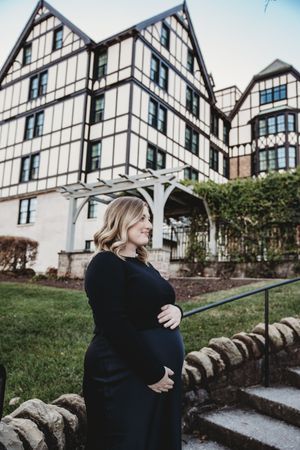 Pregnant woman in black dress cradling baby bump outdoors near Tudor-style building with stone wall.