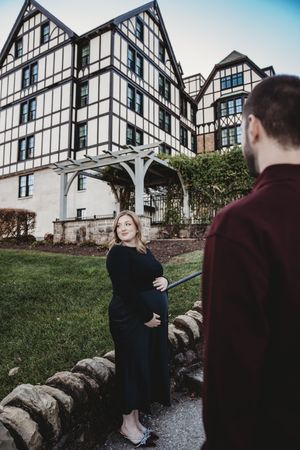 Pregnant woman in black outfit cradling baby bump outside Tudor-style building with man in burgundy shirt.