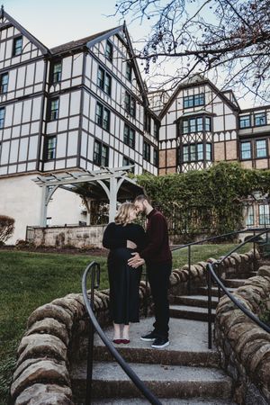 Pregnant couple sharing a tender kiss on stone steps outside a Tudor-style building during maternity photo session.