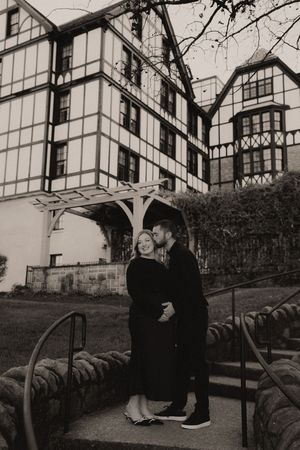 Black and white photo of a couple embracing on steps before a Tudor-style building.