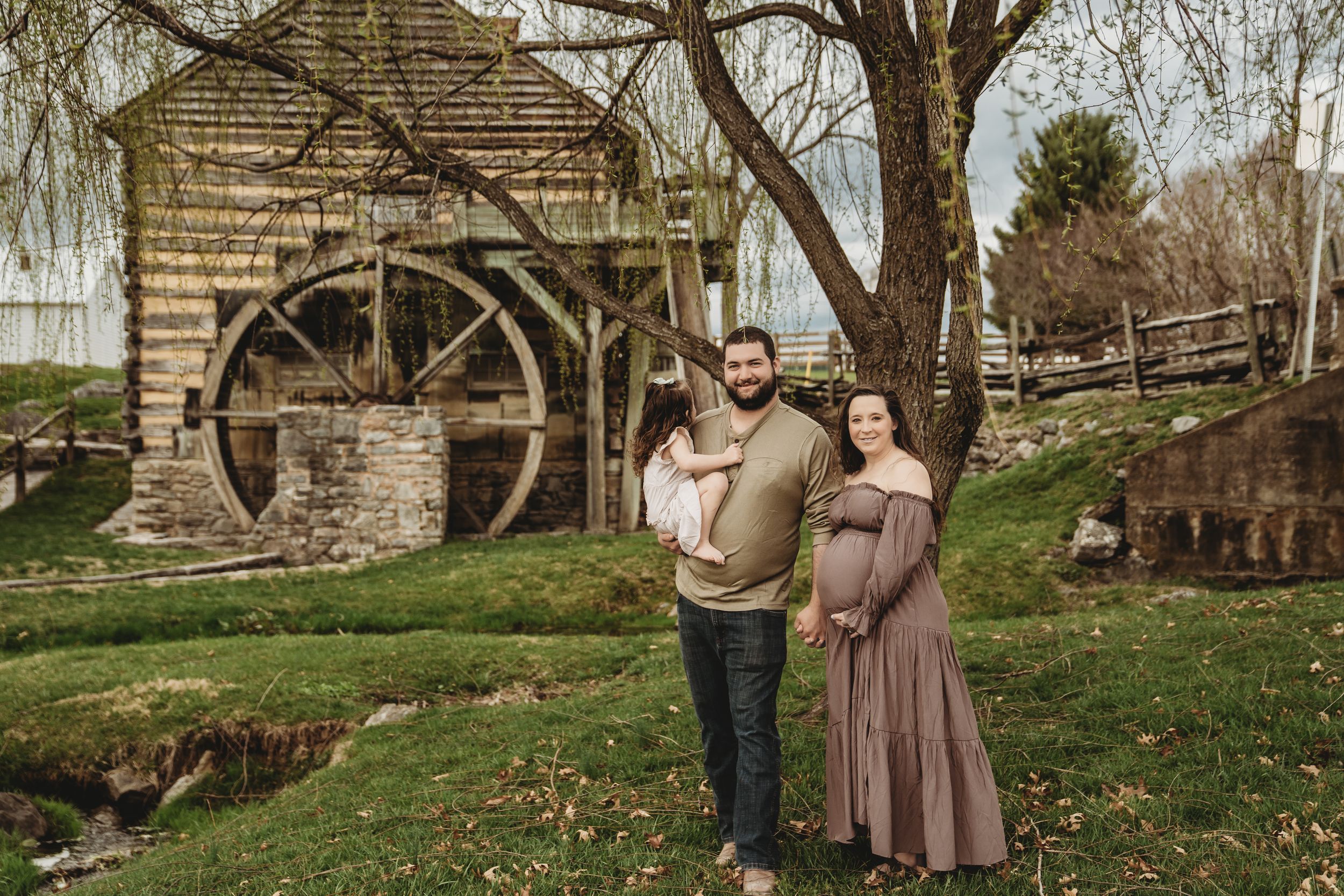 Pregnant couple with child poses before rustic grist mill with water wheel in lush green outdoor setting.