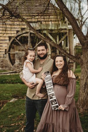 Pregnant woman holds ultrasound photos with man holding toddler girl outdoors near historic grist mill.
