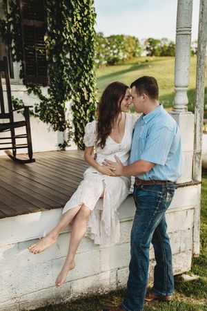 Couple sharing a romantic moment on a farmhouse porch, woman in white dress seated on railing, man in blue shirt and jeans.