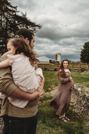 Pregnant woman in mauve dress smiles at man holding young child on a farm with stormy skies.