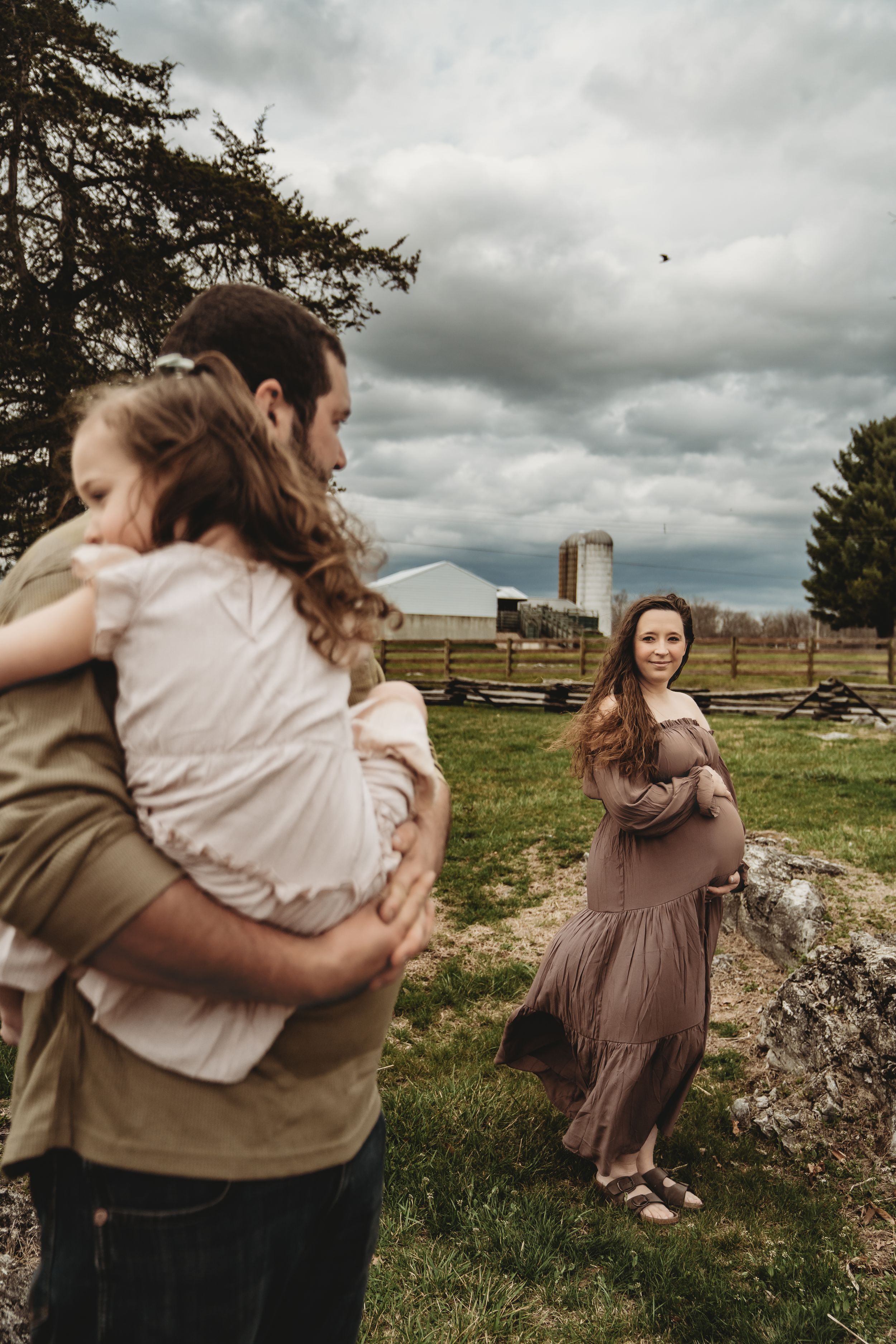Pregnant woman in mauve dress smiles at man holding young child on a farm with stormy skies.