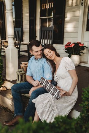Pregnant couple sitting on porch steps holding ultrasound photos, woman in white dress leaning on mans shoulder.