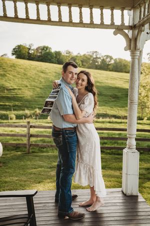 Happy couple embracing on a rustic farmhouse porch holding ultrasound photos, announcing pregnancy.