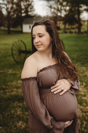 Pregnant woman in brown off-shoulder dress cradling baby bump outdoors on green grass with rustic wagon wheel backdrop.