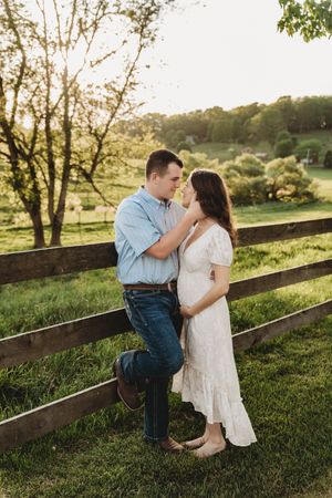 Couple shares a tender moment leaning against a wooden fence in a lush green field during golden hour.