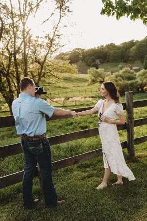 Couple holding hands by rustic wooden fence at golden hour on a lush green farm, man photographing woman in white dress.