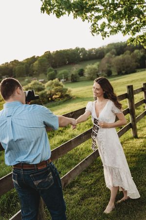 Smiling woman in white dress holds ultrasound photos while man photographs her by a wooden farm fence.