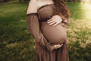 Pregnant woman in brown off-shoulder maxi dress cradling baby bump outdoors in green garden during golden hour.