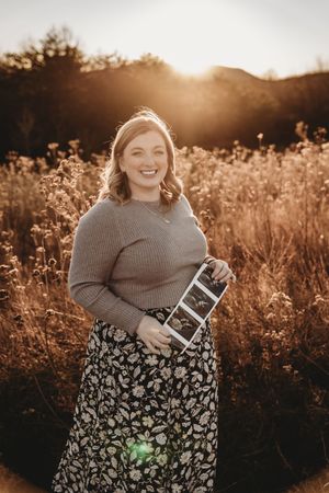 Smiling pregnant woman holding ultrasound photos in golden sunlit field wearing floral skirt and tan sweater.