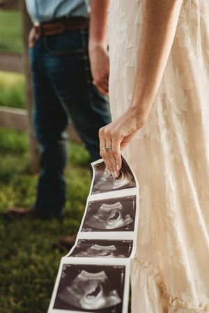 Woman in white dress holds ultrasound photos outdoors while man in jeans stands nearby during pregnancy announcement.