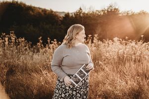 Pregnant woman holding ultrasound photos in golden sunlit field wearing gray sweater and floral skirt.