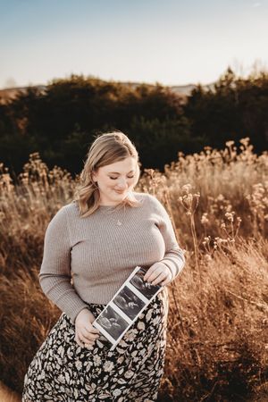Pregnant woman in floral skirt holds ultrasound photos in golden field at sunset.