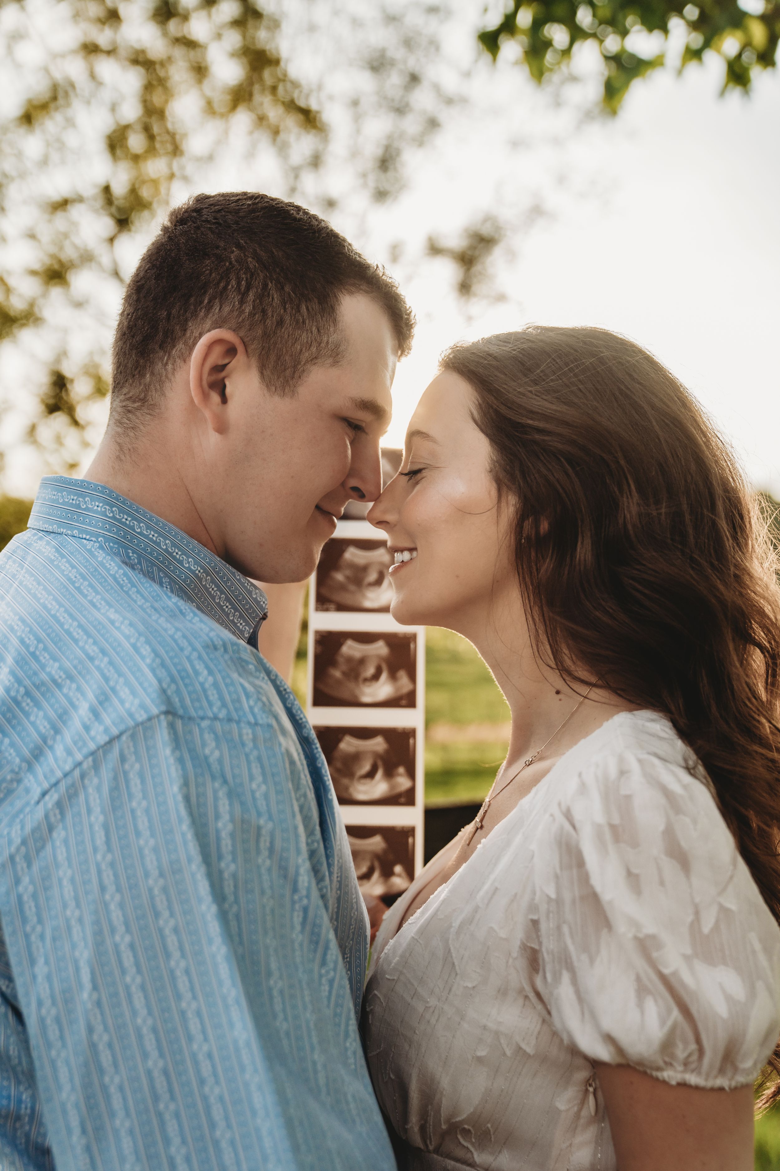A couple shares a tender nose-to-nose moment outdoors, holding ultrasound photos, announcing their pregnancy.
