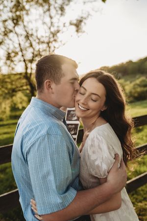 Happy couple embraces at golden hour outdoors, man kisses womans cheek as she smiles holding ultrasound photos.