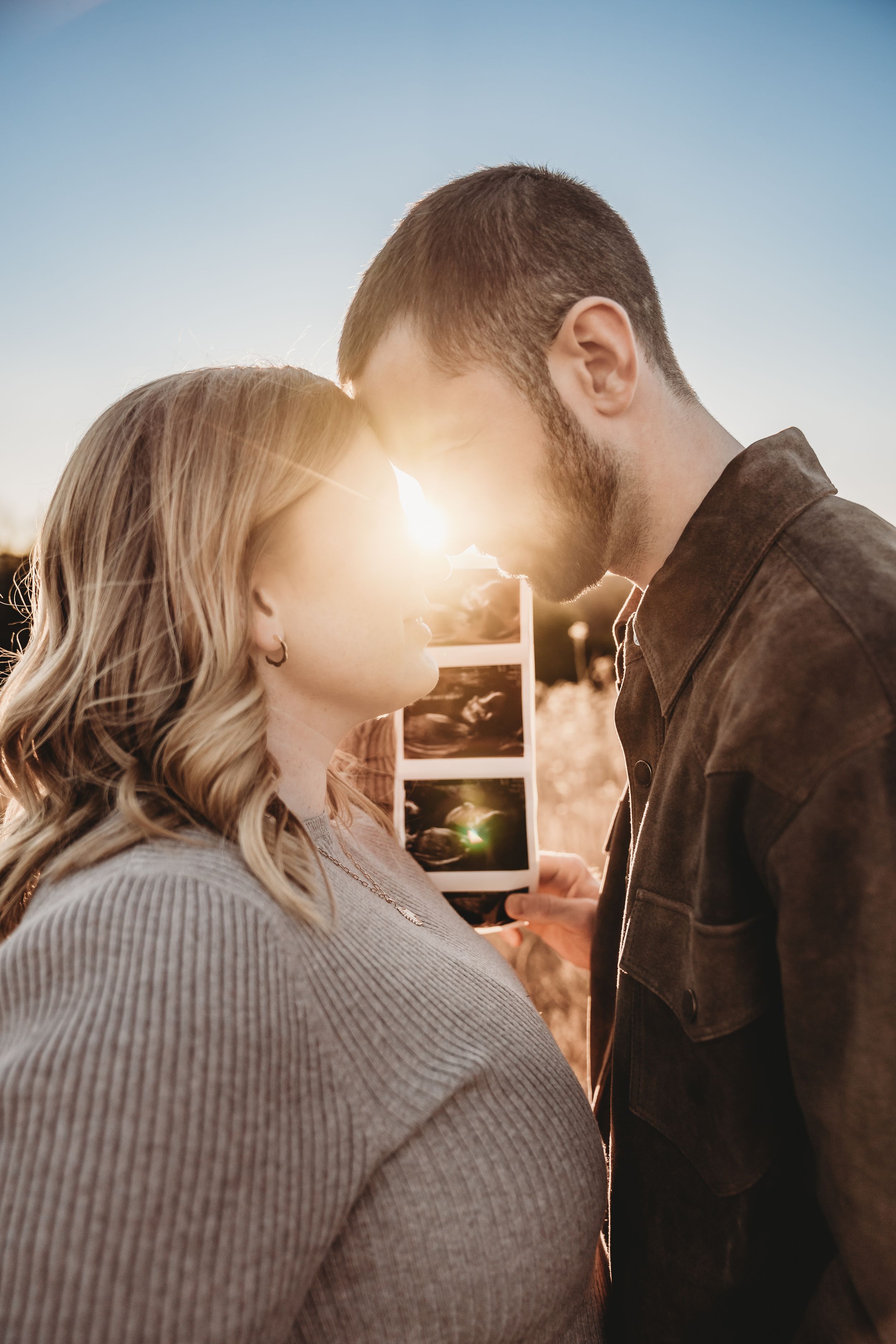 Couple touching foreheads and holding ultrasound photos backlit by golden sunset during pregnancy announcement.
