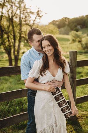 Smiling couple embracing by a wooden fence at sunset, proudly displaying ultrasound photos.
