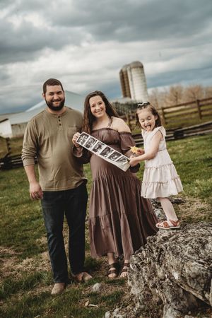 Happy family poses outdoors on a farm, holding ultrasound photos, announcing a pregnancy with their young daughter.