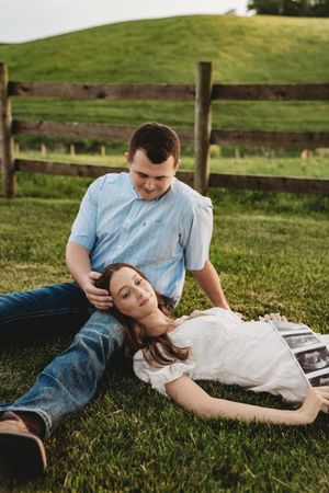 Couple relaxing on green grass near wooden fence, woman lying on man's lap holding ultrasound photos at golden hour.