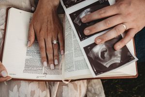 Couple's hands on open Bible with ultrasound photos, engagement ring visible, announcing pregnancy.