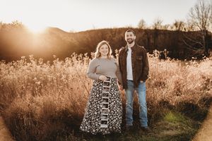 Couple stands together in sunlit field holding ultrasound photos at golden hour.